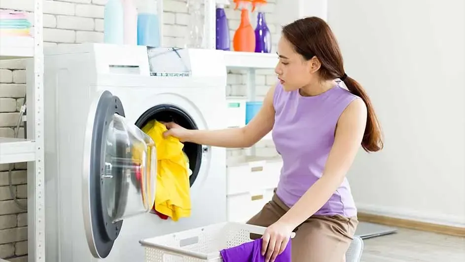 Une machine à laver blanche avec du linge jaune. Un panier blanc avec du linge violet. Des bouteilles colorées sur une étagère.
