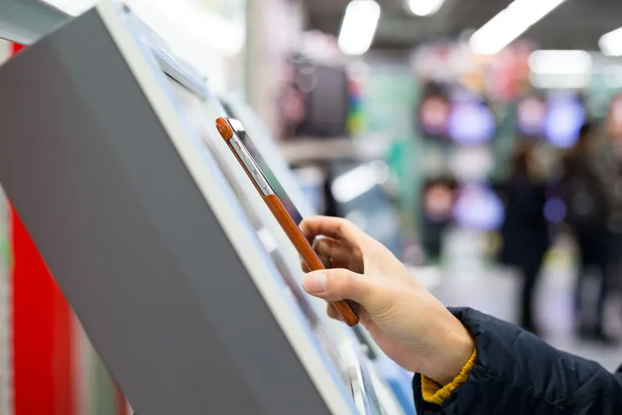 Een hand houdt een bruine smartphone vast bij een grijze interactieve kiosk in een winkel.