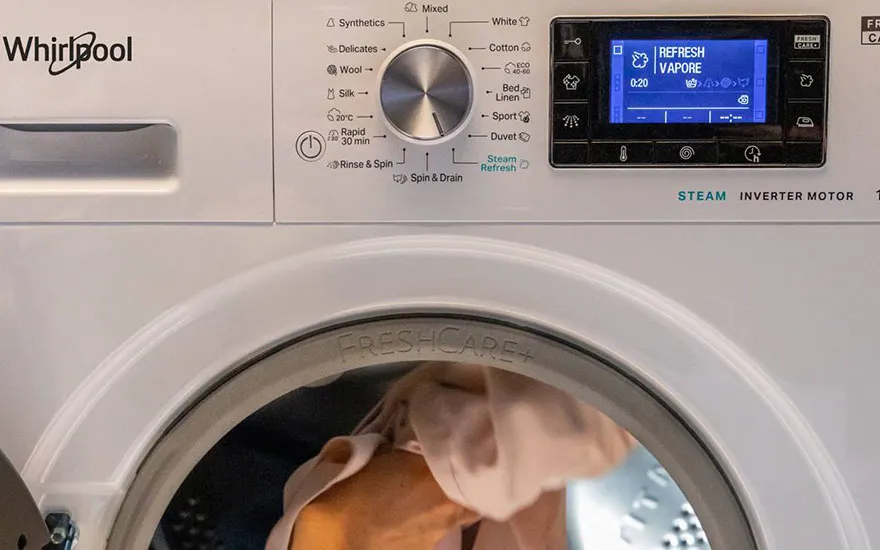Machine à laver blanche Whirlpool ouverte, avec linge à l'intérieur. Sélecteur de programme et écran LCD affichant "Refresh Vapore".