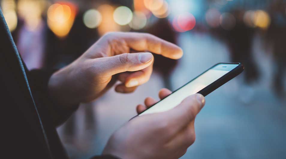 Gros plan : main tenant un smartphone allumé, doigt pointant vers l'écran. Arrière-plan flou avec des lumières.