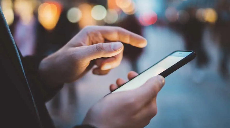 Gros plan : main tenant un smartphone allumé, doigt pointant vers l'écran. Arrière-plan flou avec des lumières.