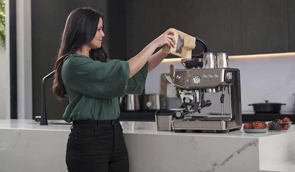 A woman in a green blouse pours coffee beans into a shiny espresso machine on a marble island, with pastries and berries neatly arranged nearby