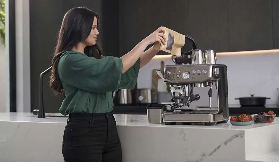 A woman in a green blouse pours coffee beans into a shiny espresso machine on a marble island, with pastries and berries neatly arranged nearby