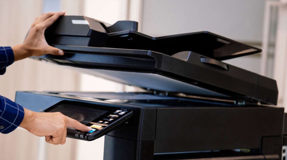 Hands operate a black office printer. One hand lifts the scanner, another presses the control panel. The background is a plain wall.