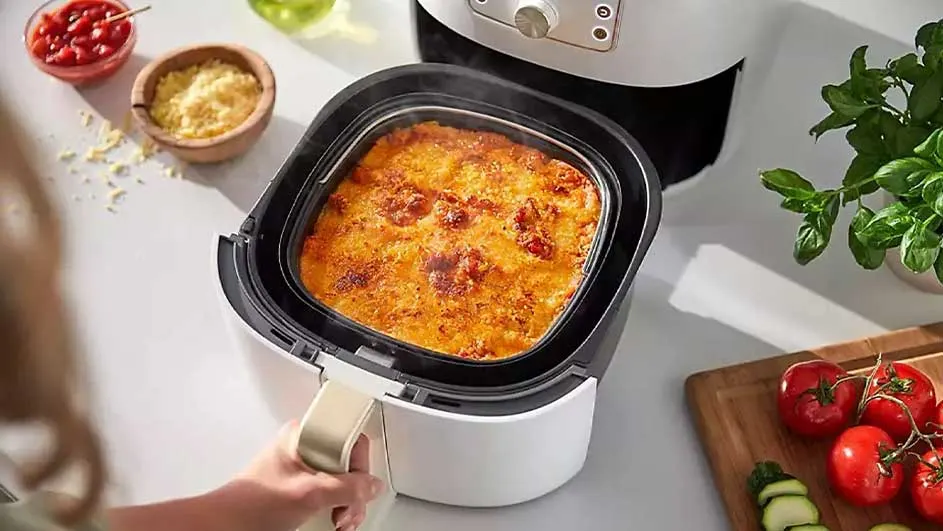 Une friteuse blanche avec un plat cuisiné à l'intérieur. A côté, des tomates, des concombres, du fromage et une sauce tomate.