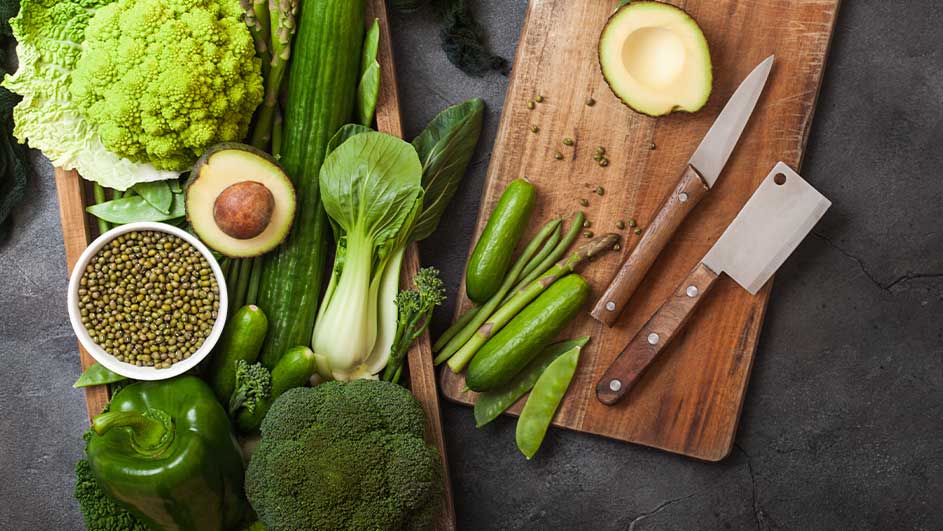 Een foto toont een houten bak en snijplank vol groenten, zoals broccoli, avocado, komkommer en groene bonen. Messen liggen op de plank.