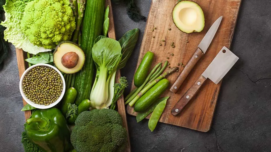 Een foto toont een houten bak en snijplank vol groenten, zoals broccoli, avocado, komkommer en groene bonen. Messen liggen op de plank.