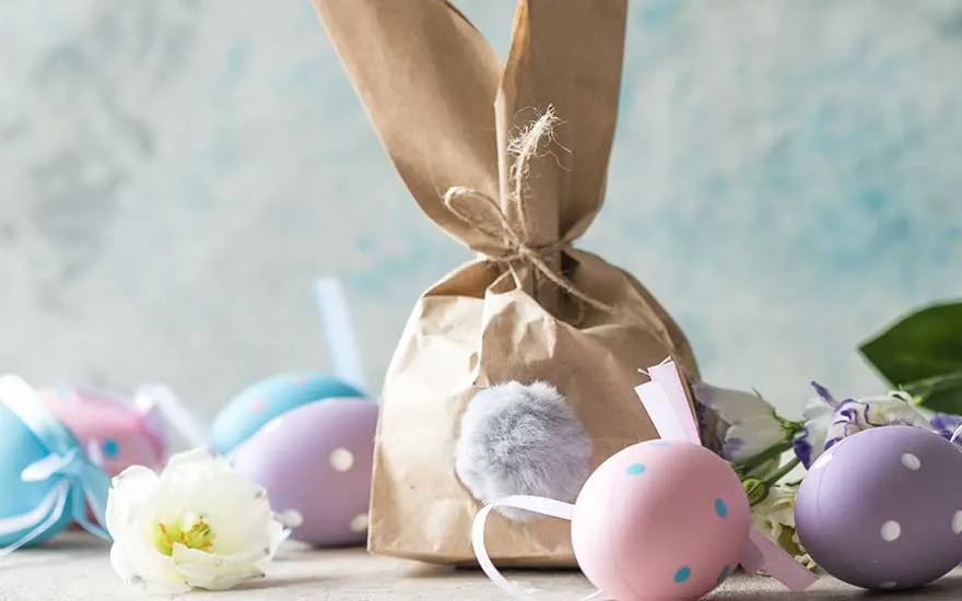 Sac en papier brun avec queue de pompon, œufs colorés à pois et fleurs sur fond texturé.