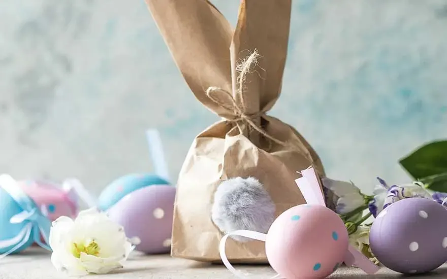 Sac en papier brun avec queue de pompon, œufs colorés à pois et fleurs sur fond texturé.