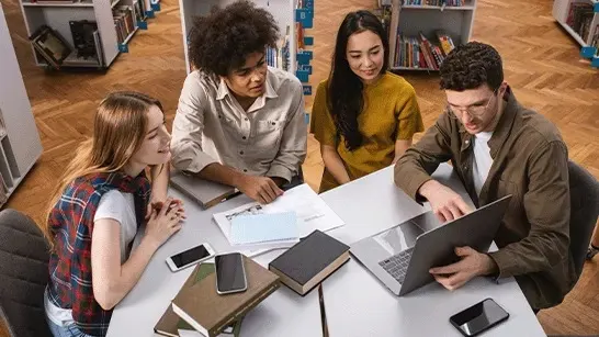 Quatre personnes sont assises autour d'une table blanche avec un ordinateur portable, des livres et des téléphones dans une bibliothèque.