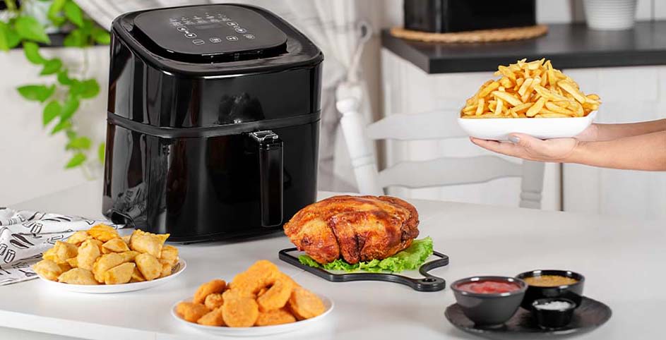 A black air fryer sits on a white table with fried food, sauces, and a hand holding fries in a white bowl.