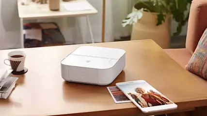 Een witte printer staat op een houten tafel, naast een telefoon en kopje koffie. Op de achtergrond staat een plant en een bijzettafel.