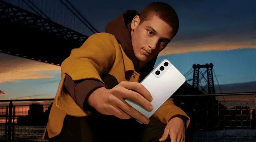 Een witte Samsung smartphone met drie camera's wordt vastgehouden tegen een achtergrond van een brug bij zonsondergang.