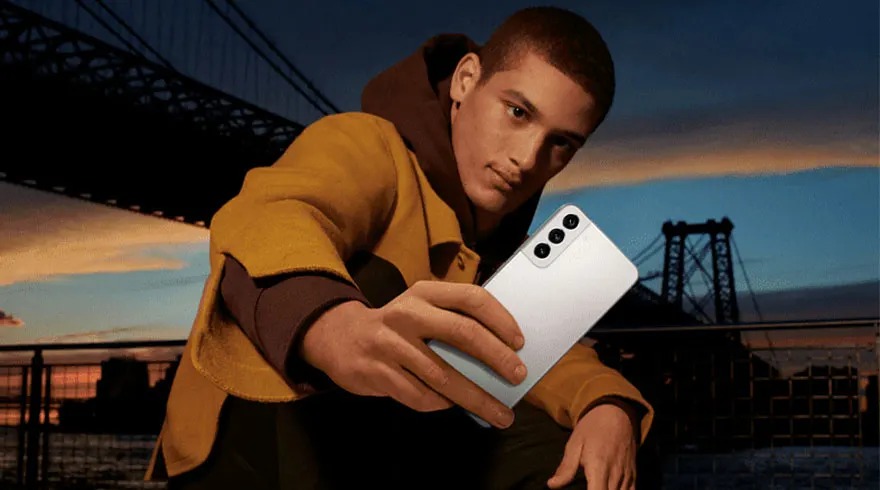 Un téléphone blanc avec trois objectifs est tenu devant un pont et un ciel au crépuscule.