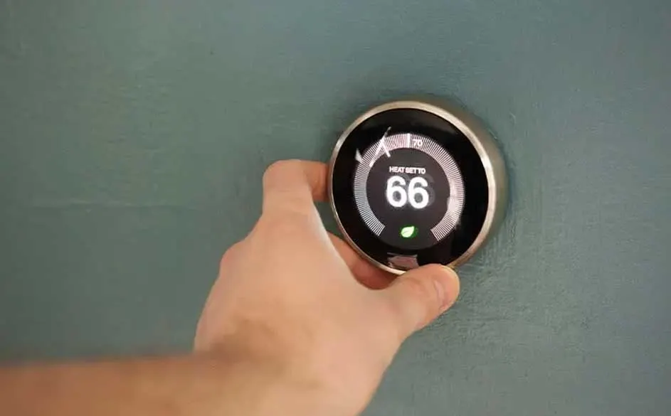 Main tenant un thermostat numérique rond fixé au mur. L'écran affiche 66 degrés. Le mur est bleu-vert.