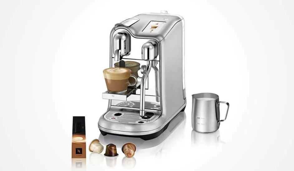 Een zilveren koffiemachine met een kop koffie, een zilveren melkkan en capsules op een witte achtergrond.
