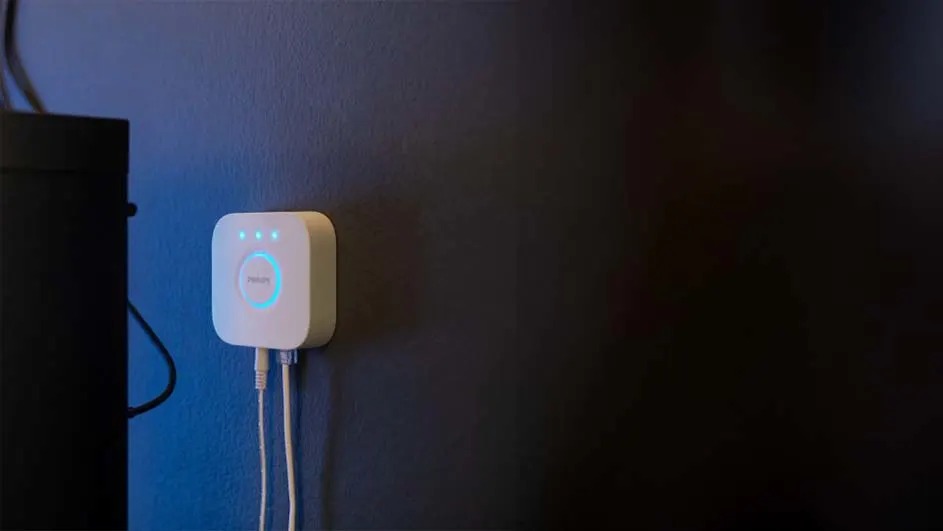 Un appareil blanc carré avec des lumières bleues est monté sur un mur bleu foncé avec des câbles blancs. Une boîte noire est à gauche.