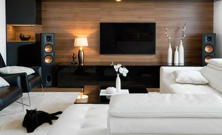 Een woonkamer met houten wand, een zwarte tv, luidsprekers, witte zetel en een zwarte kat op een wit tapijt.