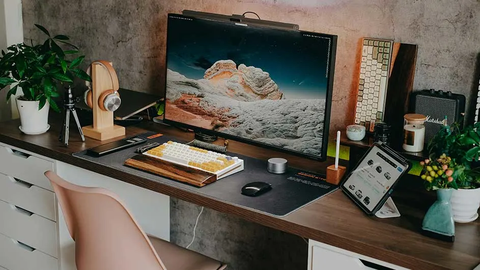 Bureau avec écran, clavier, tablette, casque audio, plante et enceinte. Le fond est un mur texturé gris.