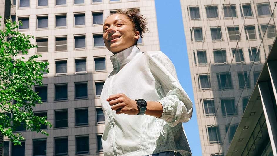 View from below of a person in a white jacket running, wearing a dark watch. Beige buildings and blue sky background.