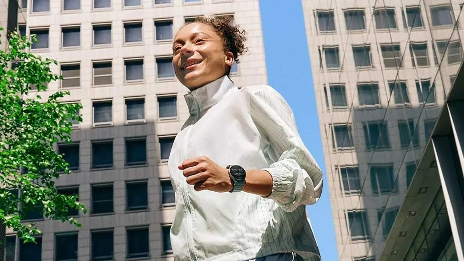 View from below of a person in a white jacket running, wearing a dark watch. Beige buildings and blue sky background.