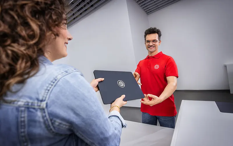 An employee in a red polo shirt receives a laptop from a customer at a service desk.