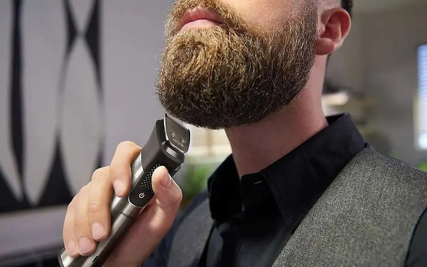 Close-up of a silver and black Philips beard trimmer cutting a light brown beard. A gray vest is visible.