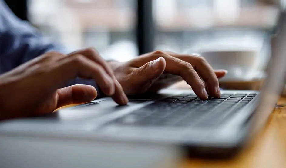 Gros plan sur deux mains qui tapent sur le clavier d’un ordinateur portable posé sur une table en bois, avec tasse de café floue près de la fenêtre.