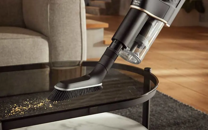 Un aspirateur noir nettoie une table basse en verre foncé avec une brosse blanche. Des miettes sont visibles.