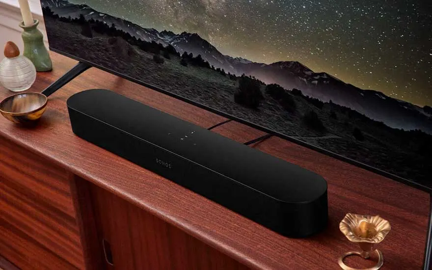 Een zwarte soundbar staat op een houten meubel onder een tv met berglandschap en sterrenhemel, omringd door elegante decoratie.