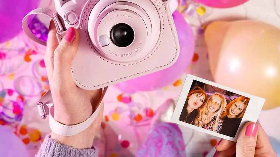 Een roze camera en foto worden vastgehouden, met ballonnen en confetti op de achtergrond.