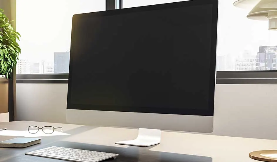 Bureau avec écran noir, clavier blanc et lunettes à côté d'une plante verte devant une fenêtre.