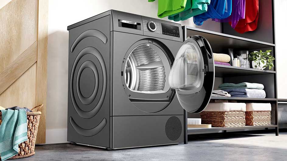 Grijze wasdroger met open deur, naast een rek met handdoeken en manden. Kleurrijke kleding hangt erboven.