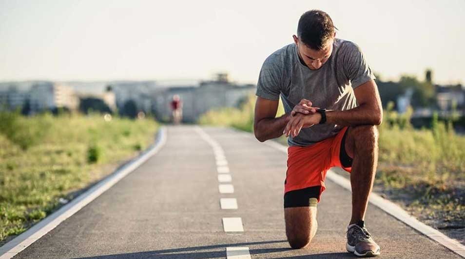 Un homme en tenue de sport vérifie sa montre sur une piste grise avec des marques blanches, herbe et horizon en arrière-plan.