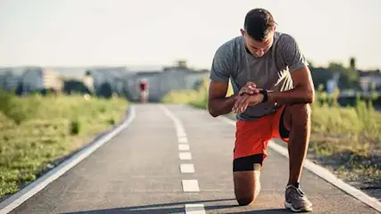 Un homme en tenue de sport vérifie sa montre sur une piste grise avec des marques blanches, herbe et horizon en arrière-plan.