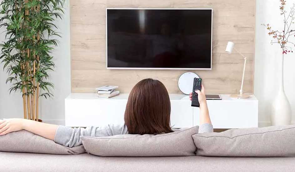 Vue arrière d'un salon avec une télé noire au mur, une commode blanche et des plantes vertes.