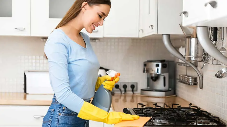 A kitchen scene shows a black stovetop being cleaned with an orange cloth and a grey bottle with a white spray nozzle.