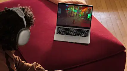 Een laptop staat op een rode bank, met een scherm dat een stadsgezicht toont. Er is ook een grijze hoofdtelefoon zichtbaar.