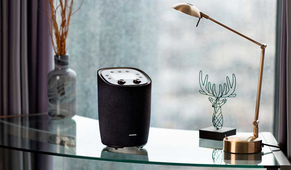 Zwarte speaker staat op een glazen tafel naast een lamp en decoratie tegen een raam met gordijnen.