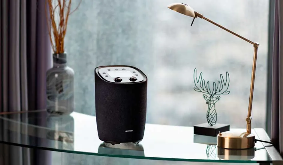 Un haut-parleur noir est posé sur une table en verre, près d'une lampe dorée et d'un vase. Un rideau est visible en arrière-plan.