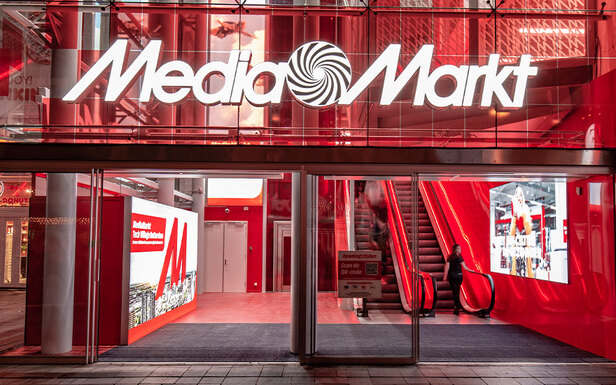 The image shows the entrance of a Media Markt store with escalators and displays visible through the glass facade. The building is red and white.