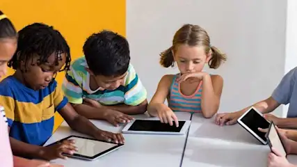 Des enfants sont assis à une table blanche, utilisant des tablettes. Le fond est jaune et blanc.