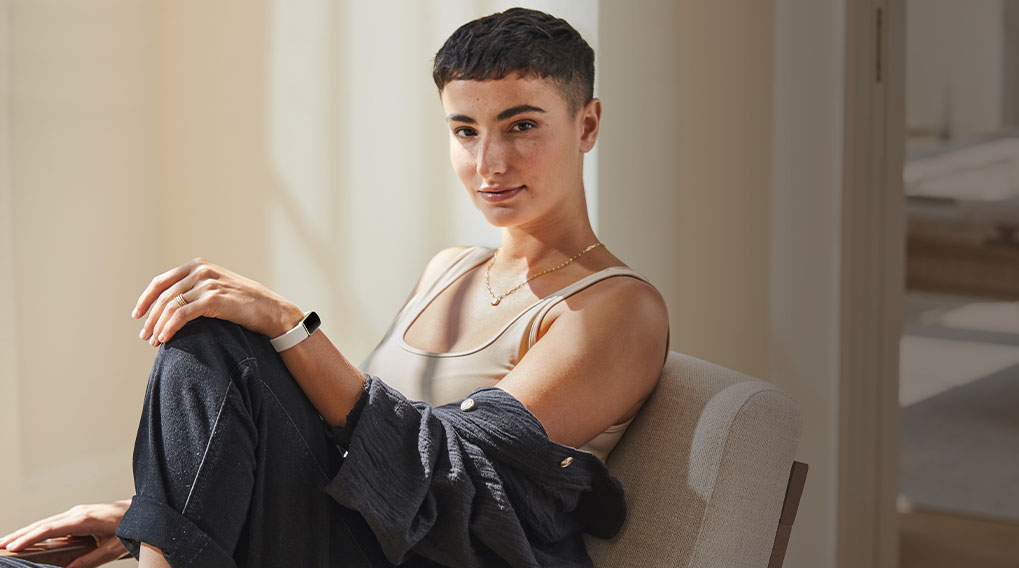 Een persoon zit op een stoel met een witte fitnesstracker om de pols. De kamer heeft zachte, neutrale kleuren.