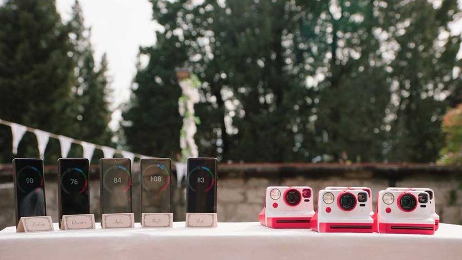 Vijf telefoons en drie roze camera's op een witte tafel. Bomen en witte vlaggetjes op de achtergrond.