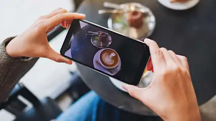 Une personne prend en photo un cappuccino et un dessert sur la table avec son smartphone, vue de dessus.