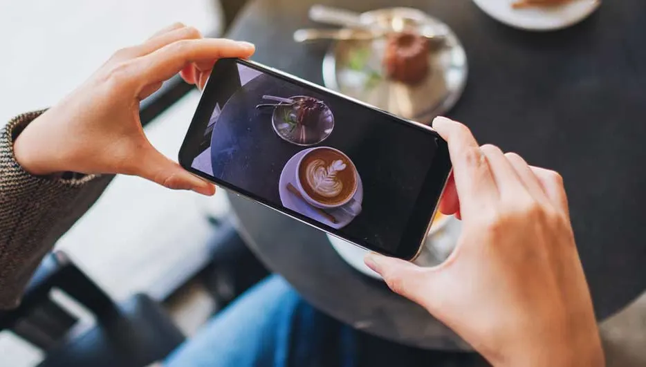 Une personne prend en photo un cappuccino et un dessert sur la table avec son smartphone, vue de dessus.