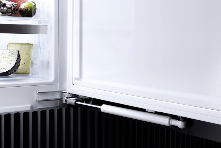 Un frigo blanc ouvert montre du fromage, des fruits. Une porte blanche est fixée avec un piston blanc sur un bord noir.