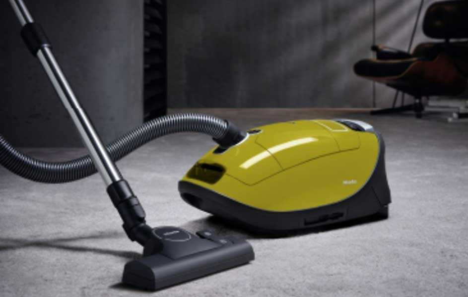 Un aspirateur traîneau jaune et noir avec tube et brosse sur un sol gris, devant un mur gris et un fauteuil.