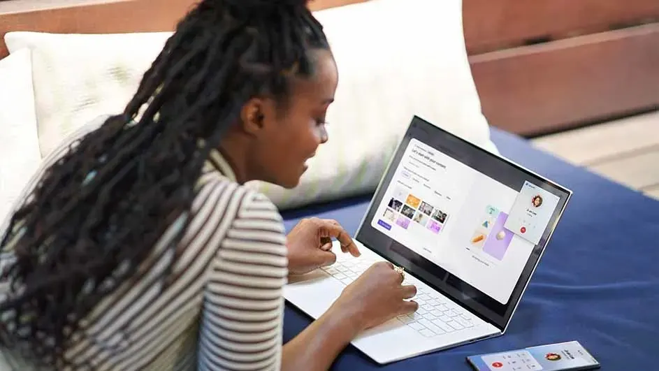 Een witte laptop met een website op een blauwe ondergrond, een witte kussen achter de laptop. Een gsm ligt naast de laptop.
