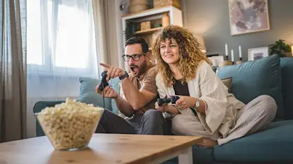 Twee personen zitten op een blauwe bank met controllers. Op de tafel staat een kom popcorn. Interieur van een woonkamer.